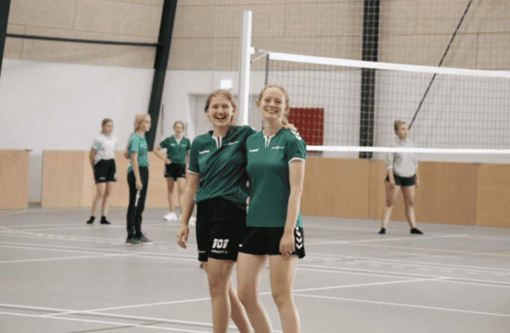 To meget smilende piger i skolens sportshal med volleyballspillere i baggrunden.